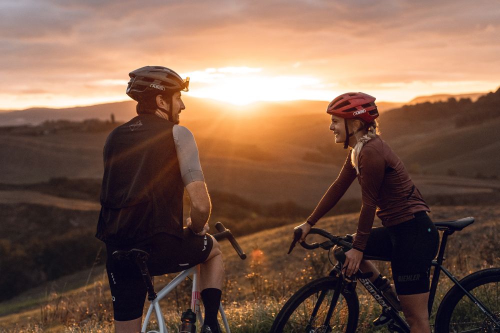 2 Personen mit Rad in hügeliger Landschaft beim Sonnenuntergang